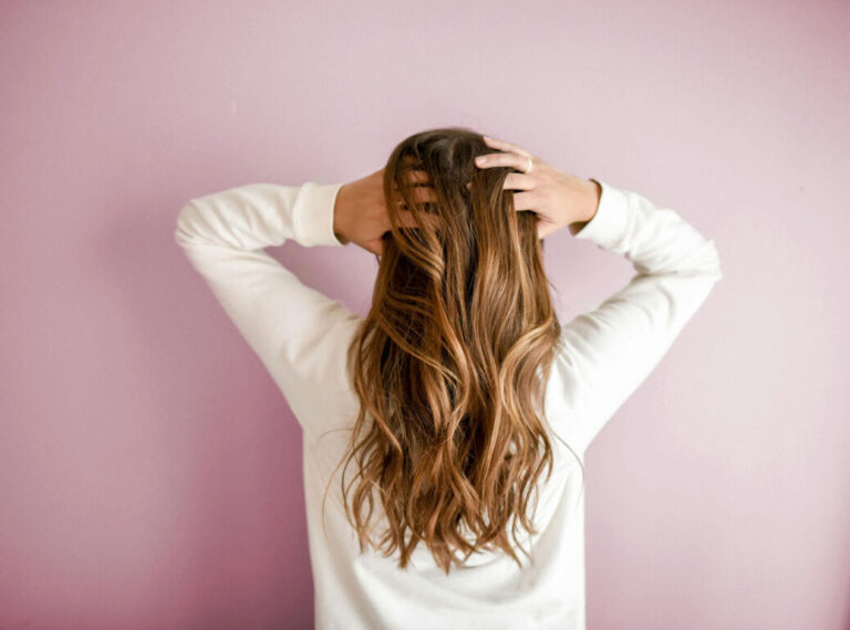 Woman with long, wavy light brown hair, seen from behind, holding her head with both hands against a soft pink wall. She is wearing a white long-sleeve top.