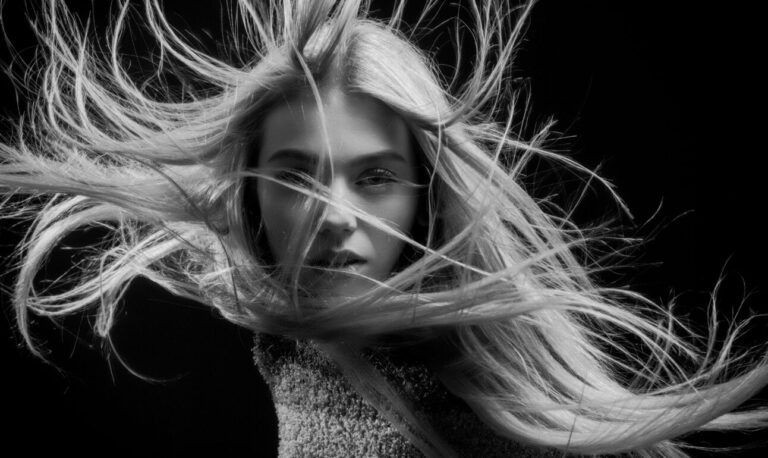 Black and white portrait of a woman with long, flowing hair blowing in different directions against a dark background.
