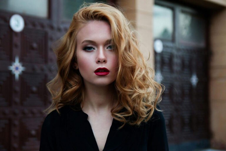 Confident woman with voluminous wavy blonde hair and bold red lipstick wearing a black top, standing outdoors in front of ornate wooden doors