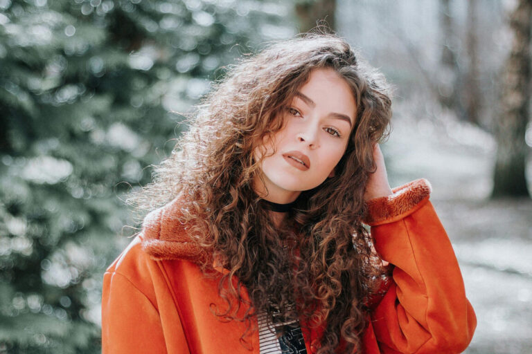 Woman with long curly hair wearing an orange coat with a fur collar, posing outdoors in soft natural light with trees blurred in the background