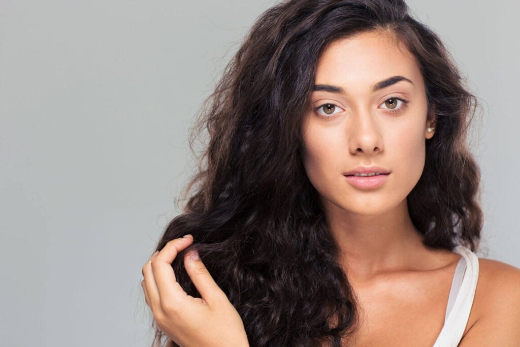 Beauty Blog Young woman with long, dark, naturally curly hair touching a strand and wondering how to repair damaged hair fast.