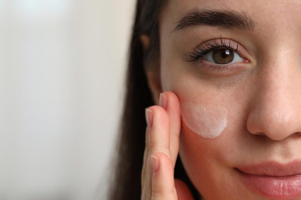 Beauty Blog Young woman with dry skin applying cream onto her face on blurred background, closeup