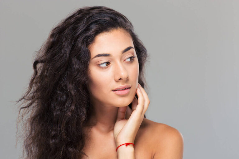 Woman with long wavy dark hair and natural makeup looking to the side, touching her face gently against a neutral gray background.