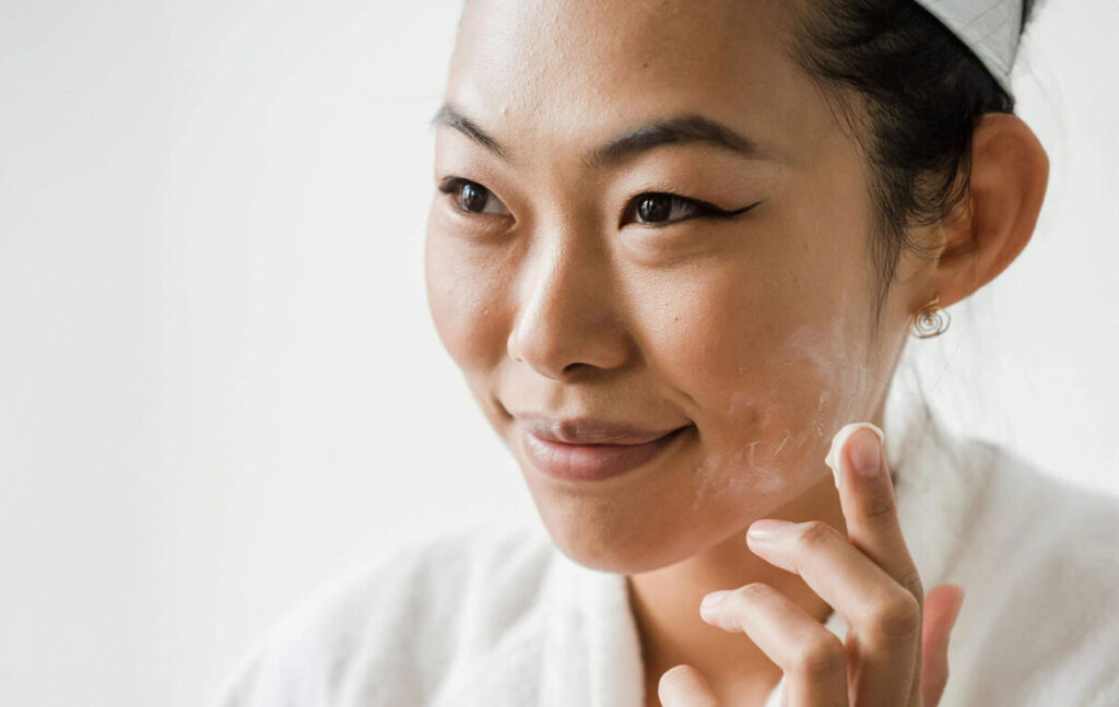 Beauty Blog A woman wearing a white robe and headband applies face cream to her cheek while smiling, holding a small jar of moisturizer in her other hand.