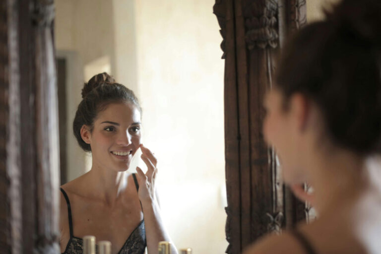 A woman with her hair in a bun smiles at herself in a mirror while applying skincare
